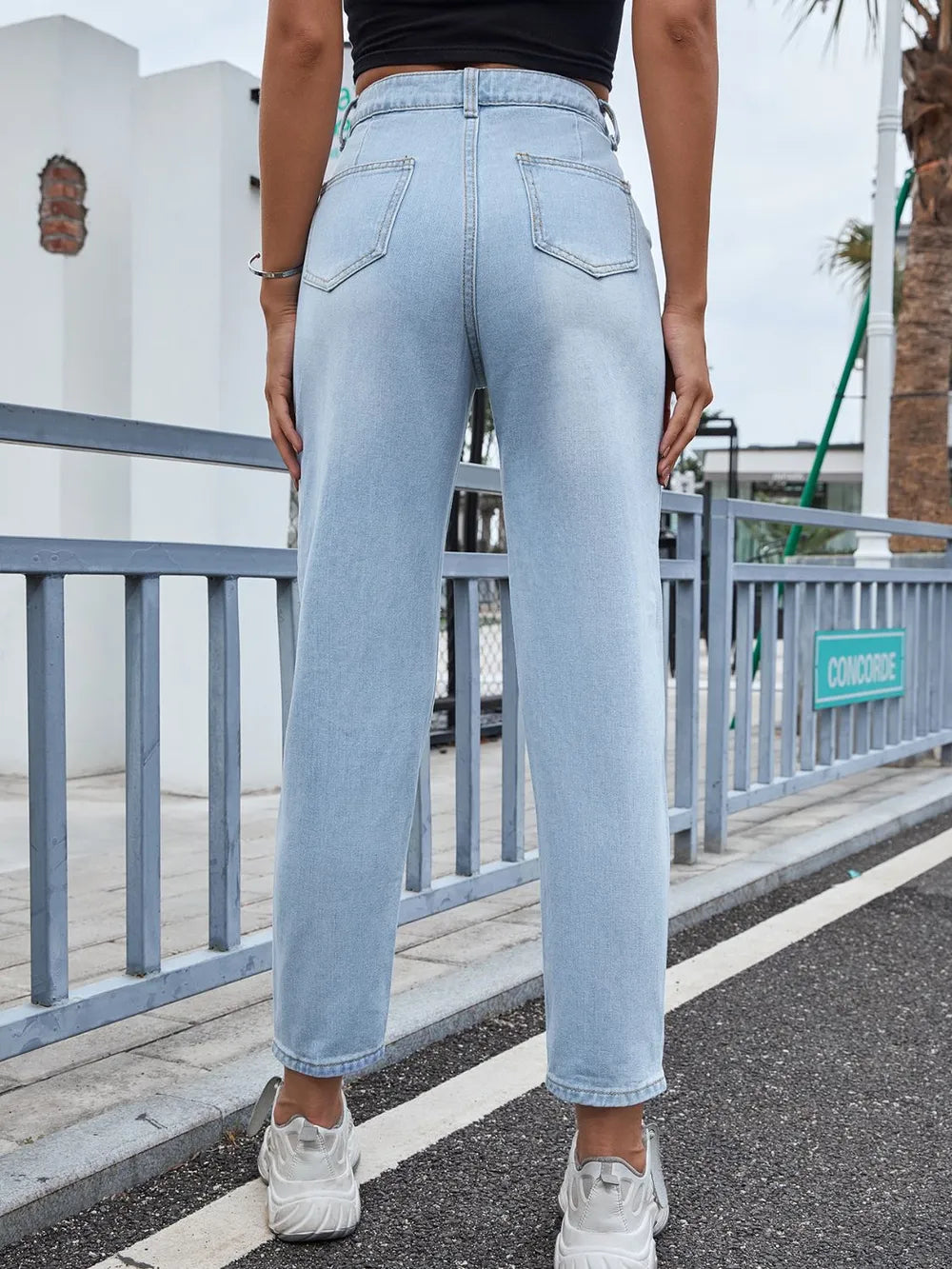 Person wearing light blue jeans standing on a street with a metal railing and building in the background.