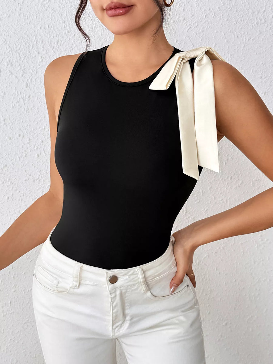 Person wearing a black sleeveless top with a white bow and white pants against a light background