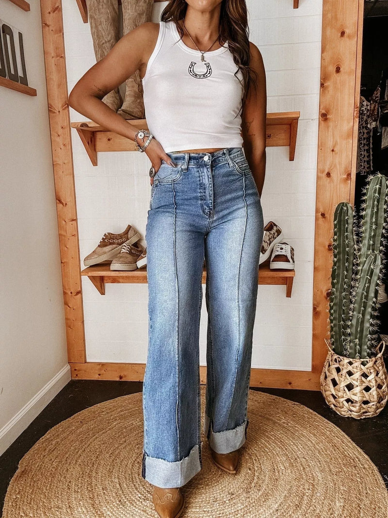 Person wearing a white tank top and blue jeans standing in a room with wooden shelves and a cactus plant.