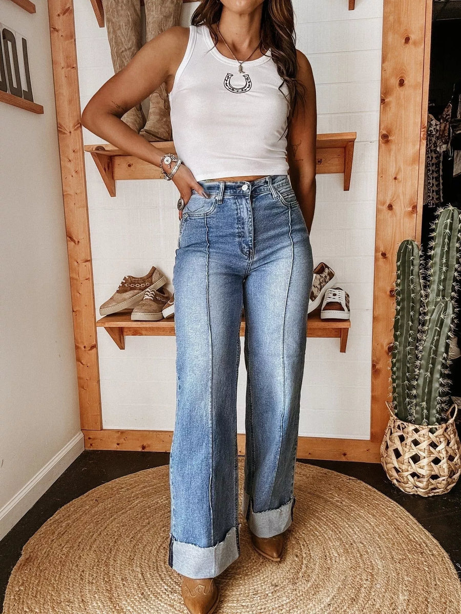 Person wearing a white tank top and blue jeans standing in a room with wooden shelves and a cactus plant.