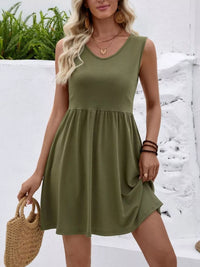 Woman wearing a green dress holding a straw bag against a white wall with greenery.