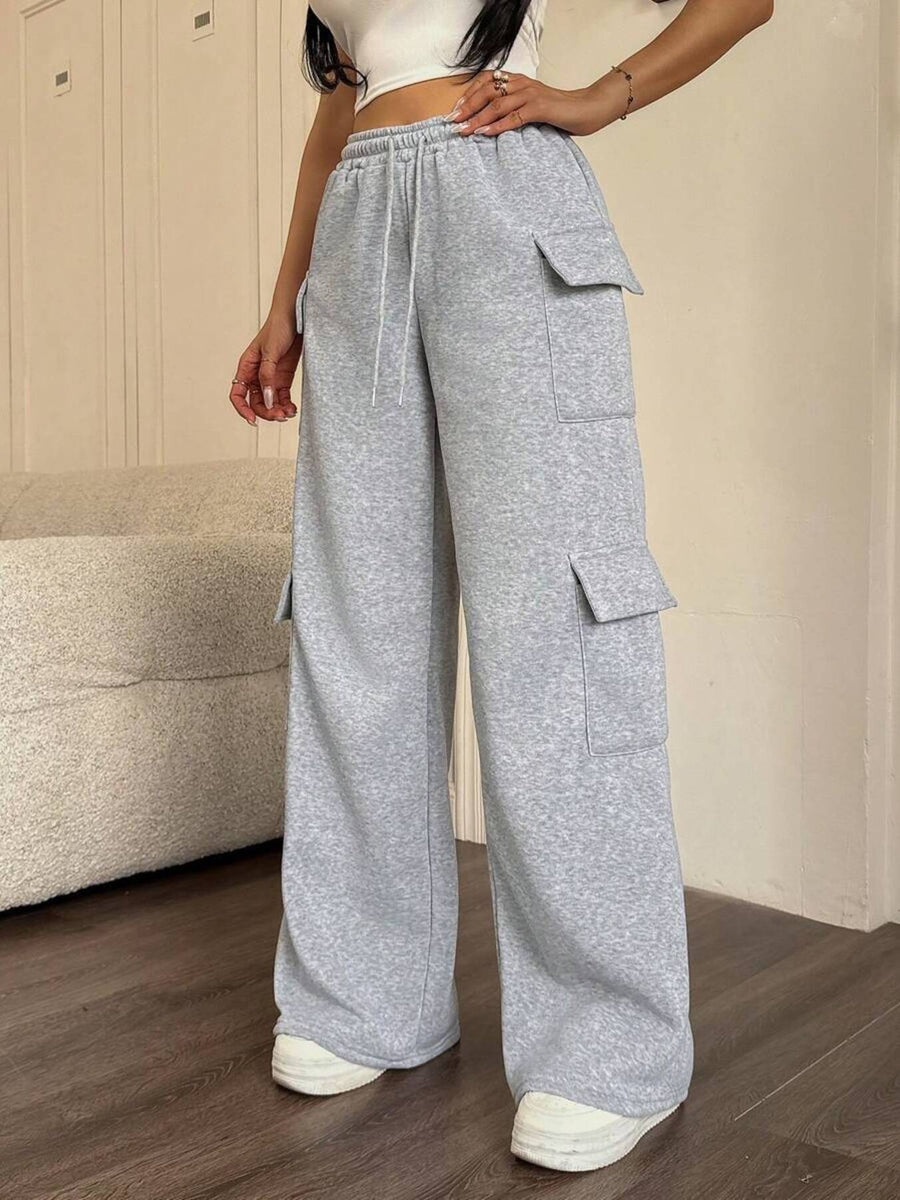 Person wearing gray cargo pants standing in a room with wooden flooring and a beige wall.