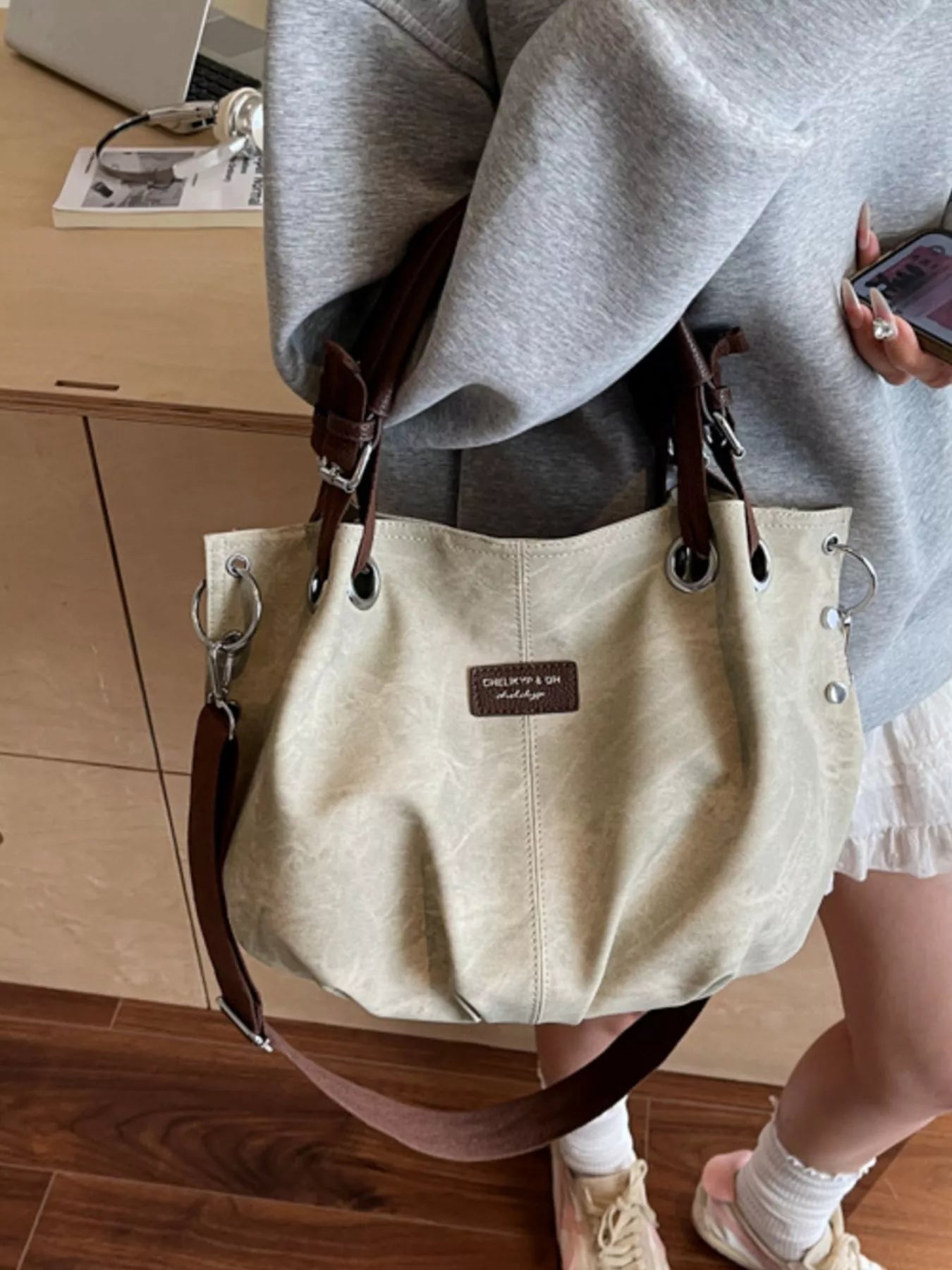 Beige handbag with brown straps worn by a person on a wooden floor.