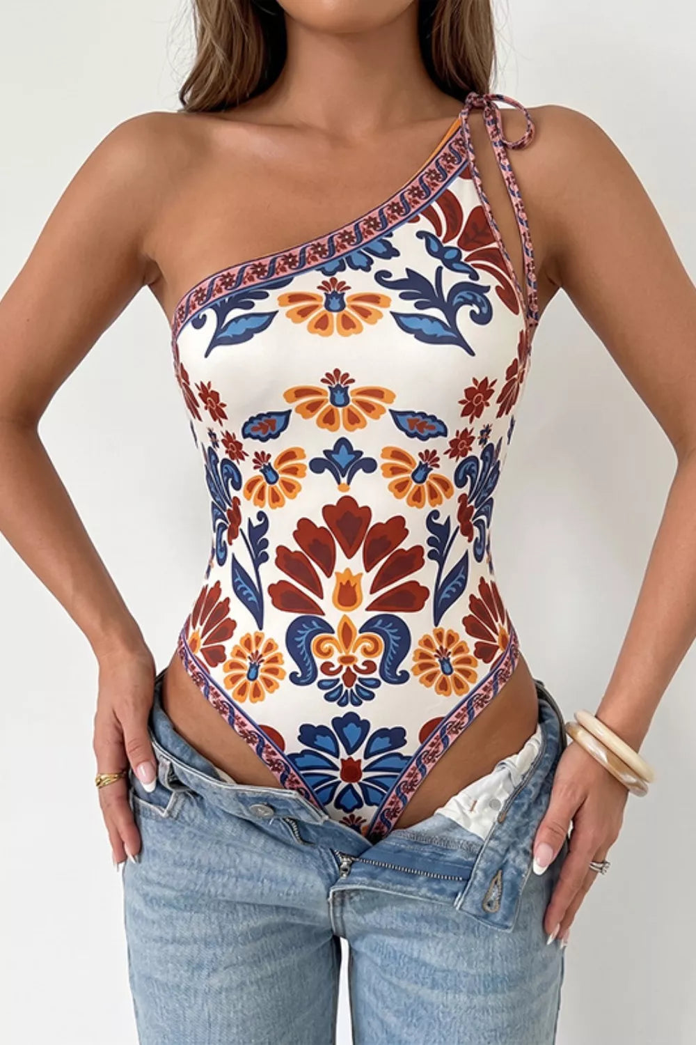 Woman wearing a patterned one-shoulder top with floral design and denim pants.