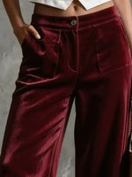 A close-up of a person wearing high-waisted, wide-leg trousers in a deep burgundy velvet fabric. The pants feature a button closure, front pockets, and a satin-like sheen, shown against a neutral gray background.