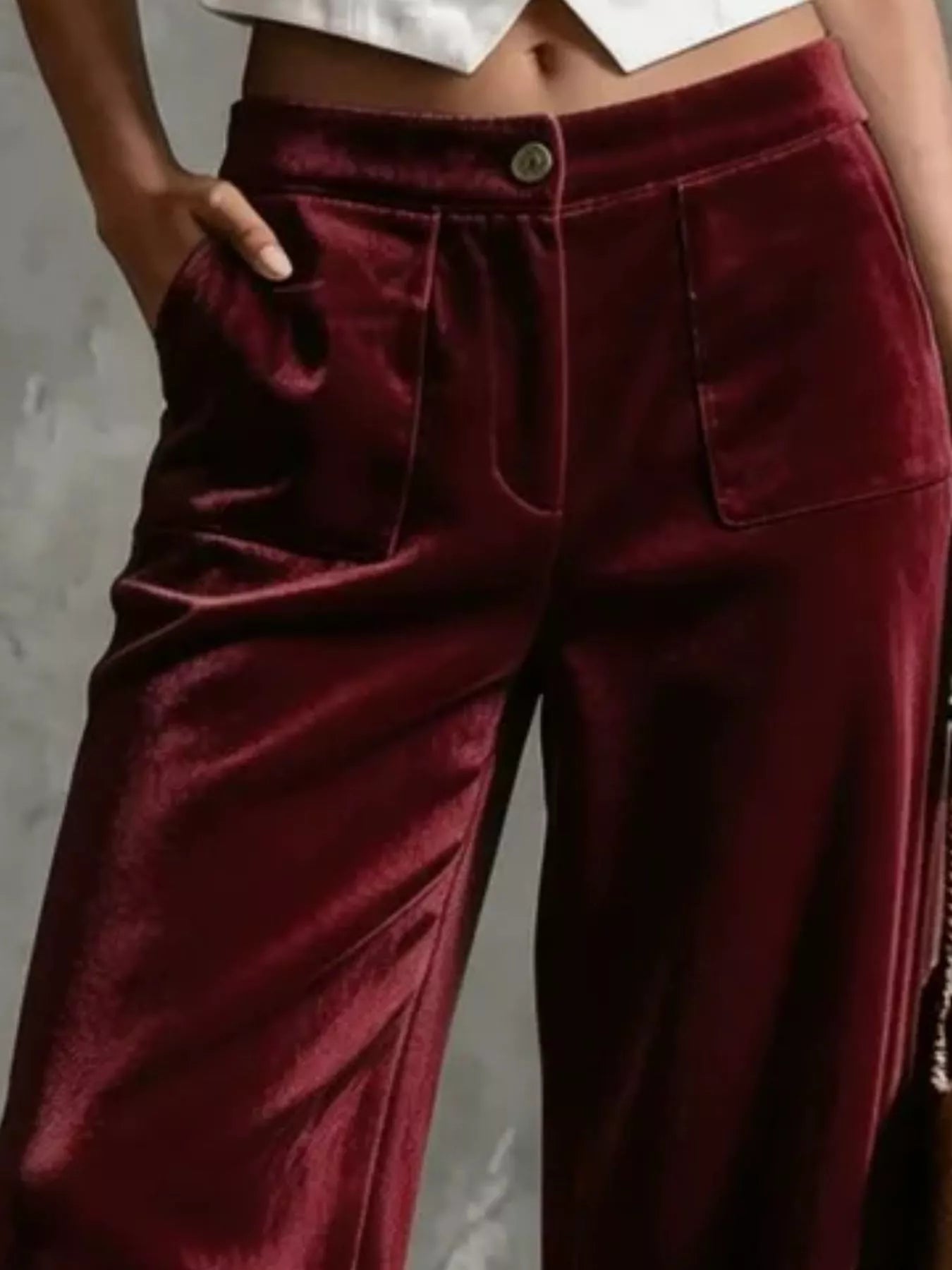 A close-up of a person wearing high-waisted, wide-leg trousers in a deep burgundy velvet fabric. The pants feature a button closure, front pockets, and a satin-like sheen, shown against a neutral gray background.