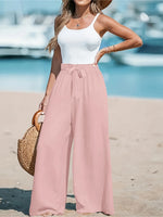 Woman in white top and pink pants standing on a beach with boats in the background