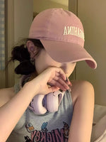 Person wearing a pink cap with text, holding pink headphones, in a casual setting.