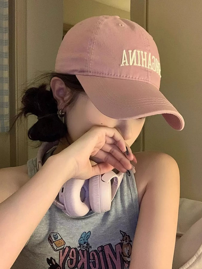 Person wearing a pink cap with text, holding pink headphones, in a casual setting.