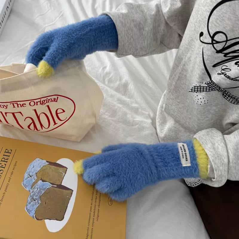 Person wearing blue fuzzy gloves with yellow cuffs, sitting on a white surface with a book and bag.