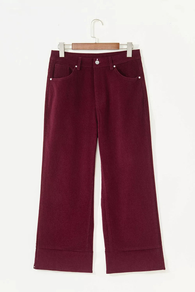 A pair of dark burgundy cropped pants with a wide leg and cuffed hems, hanging on a wooden hanger against a plain white background.