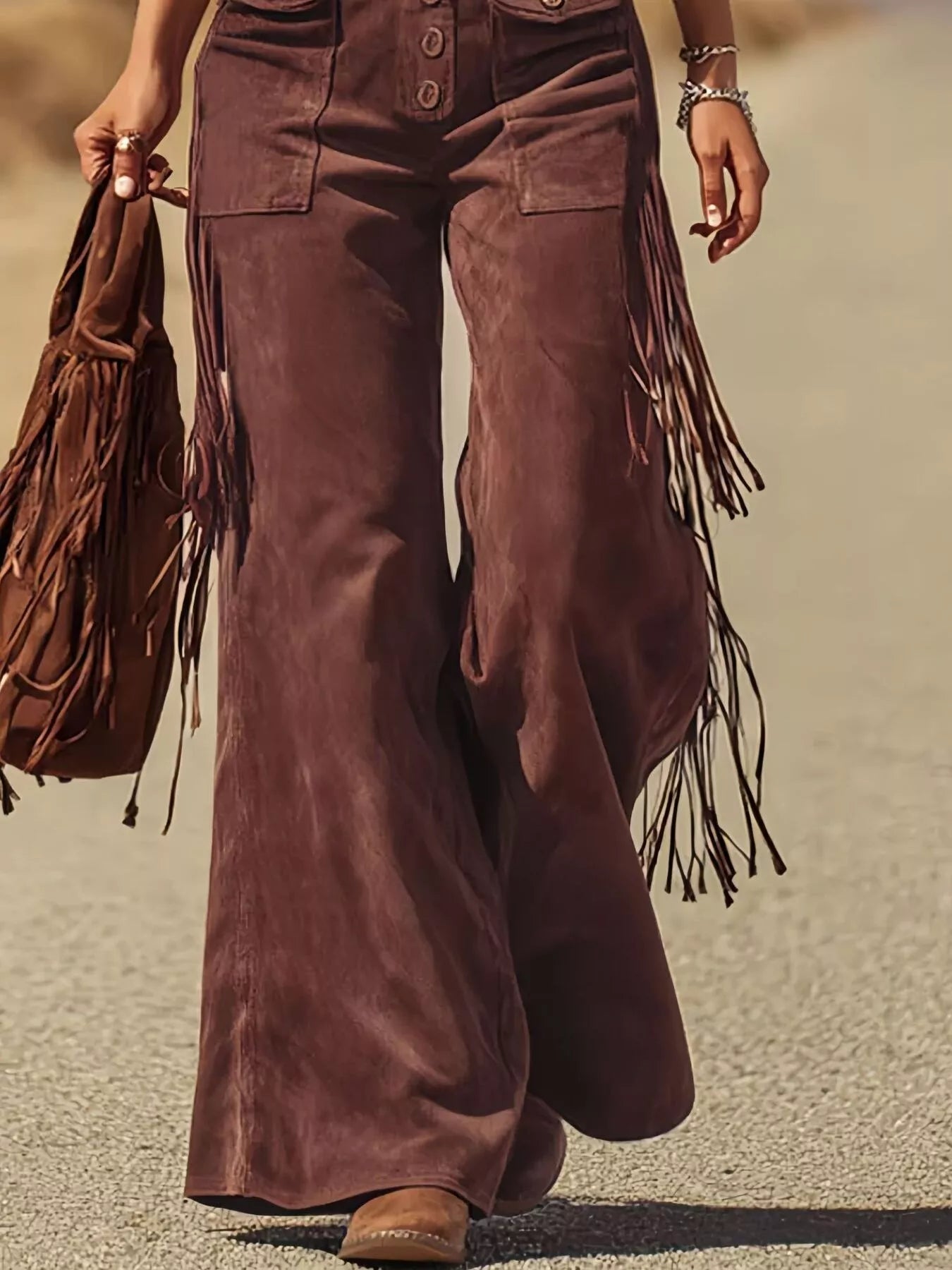 Brown suede pants with fringe details worn by a person, holding a matching brown fringed bag.