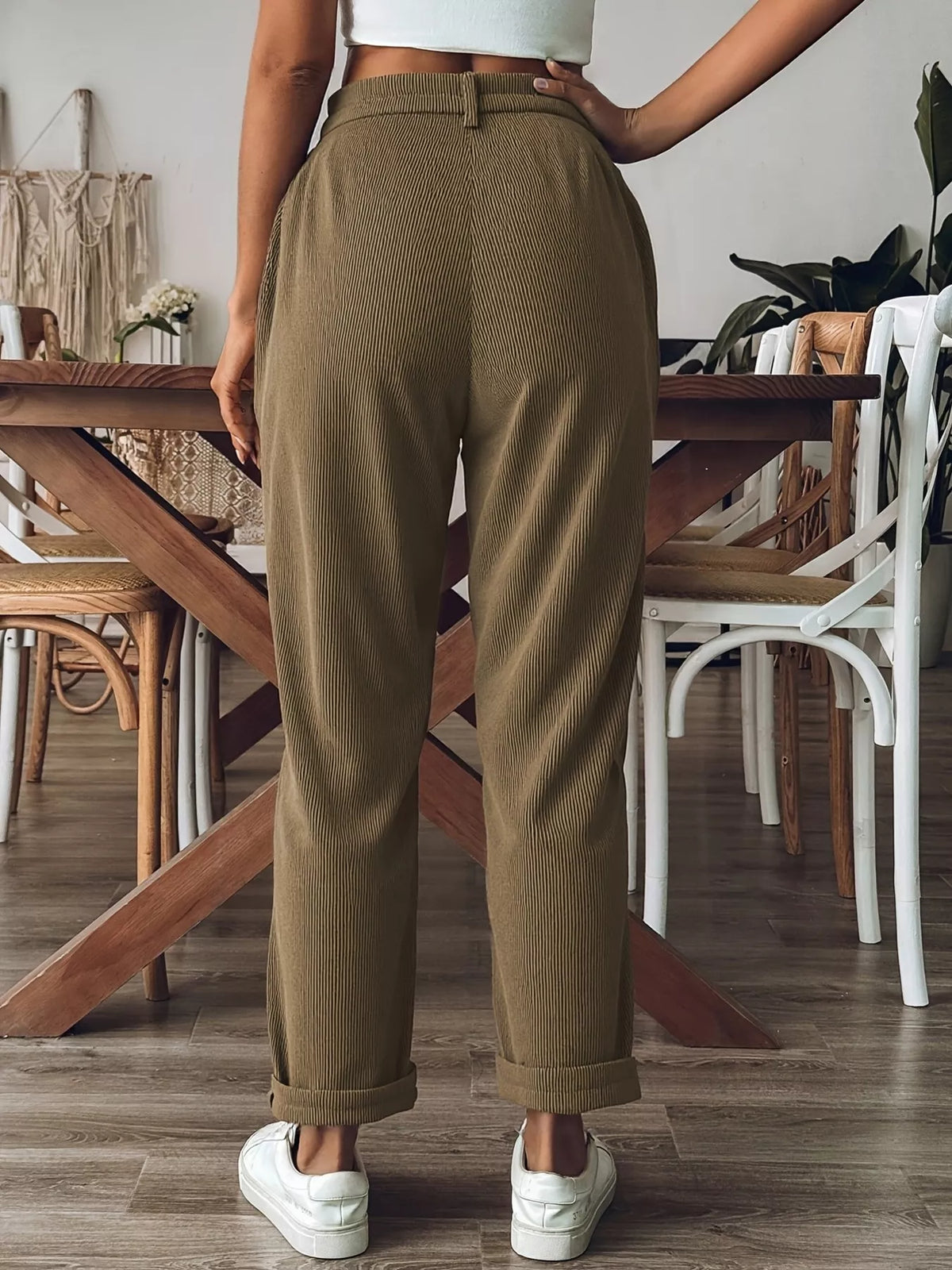 A person shown from the back wearing high-waisted, wide-leg brown corduroy trousers with cuffed hems, paired with white sneakers in a modern dining room setting.