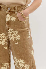 Close-up of a person wearing tan denim jeans with a cream floral print, showing the front pocket area, waistband, and hand in the pocket. A gold beaded bracelet is also visible on the wrist against a plain light background.