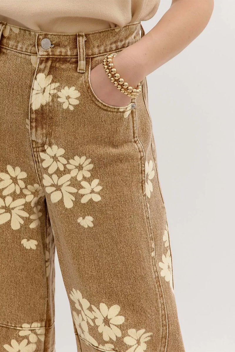 Close-up of a person wearing tan denim jeans with a cream floral print, showing the front pocket area, waistband, and hand in the pocket. A gold beaded bracelet is also visible on the wrist against a plain light background.