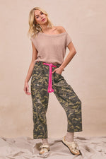 Woman wearing a beige off-shoulder top and camouflage pants with a pink belt against a beige background.