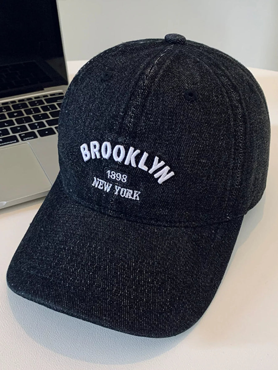 Black cap with 'Brooklyn 1898 New York' text on a light surface with a laptop in the background.