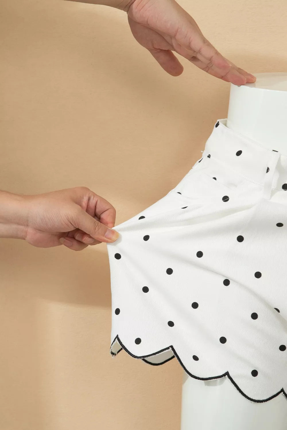White polka dot skirt with scalloped hem being held by hands against a beige background