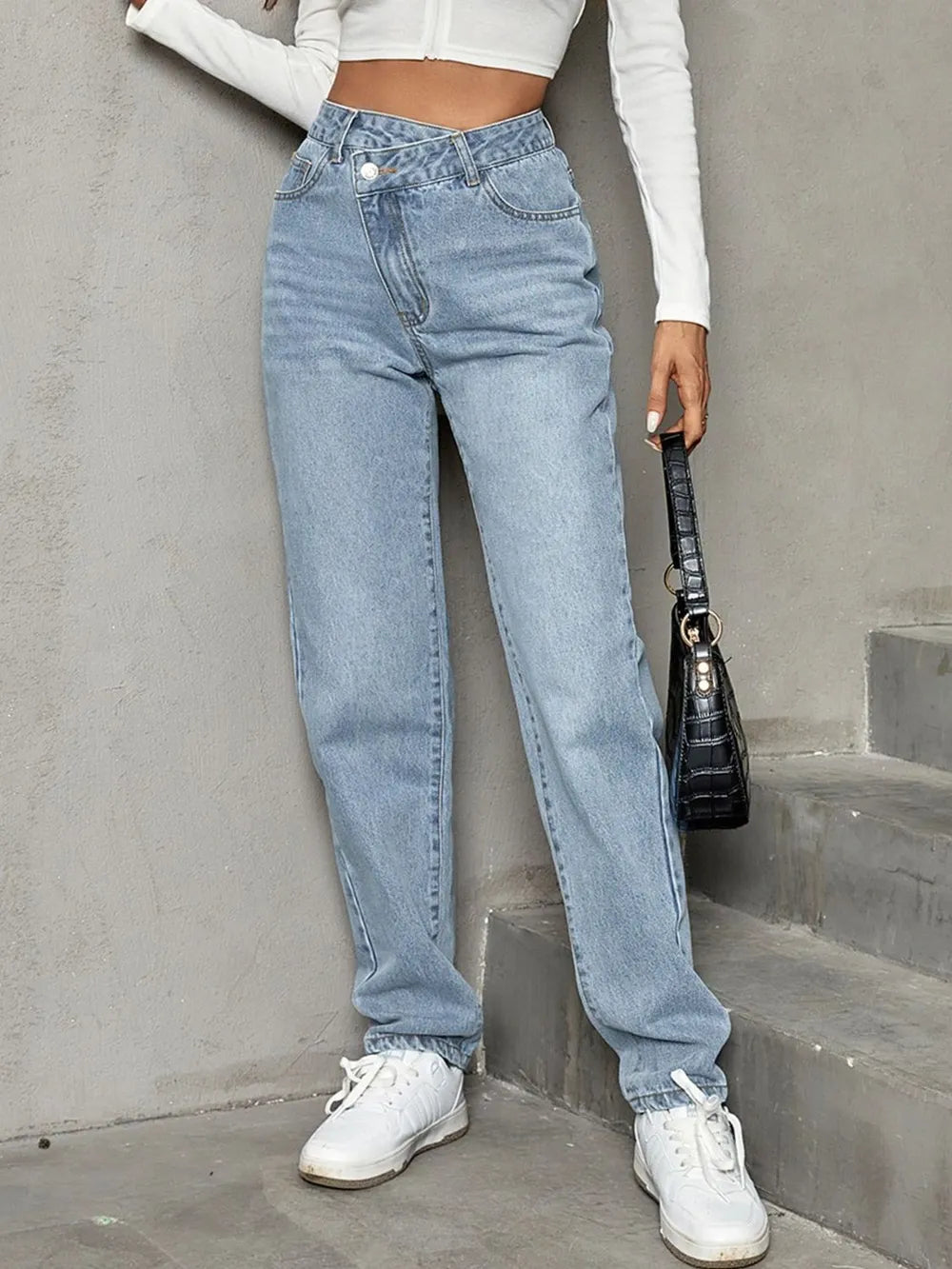 Person wearing light blue jeans and white sneakers against a gray concrete wall.