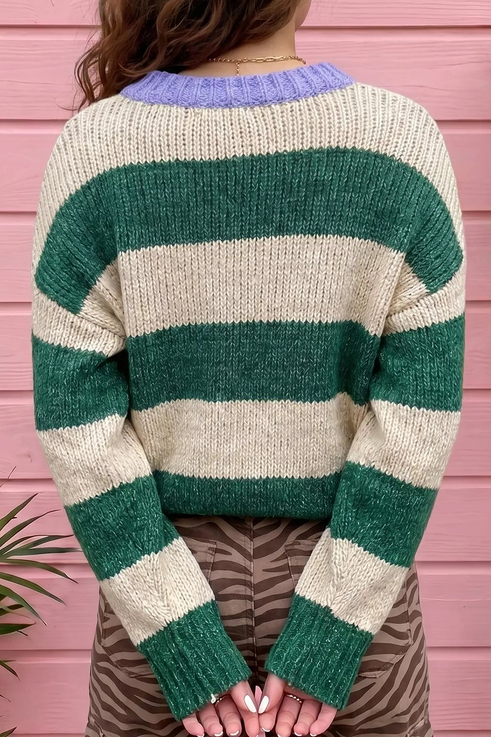 A person shown from the back wearing a chunky knit sweater with wide horizontal stripes in forest green and cream, featuring a light purple ribbed crew neck. The background consists of pink wooden siding.