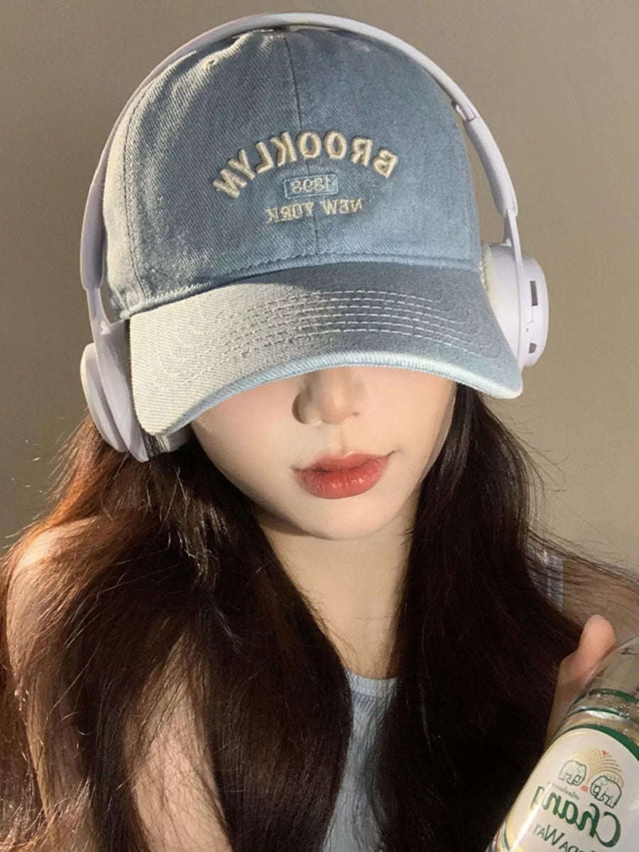 Person wearing a denim cap with 'Brooklyn' text and white headphones, holding a can.