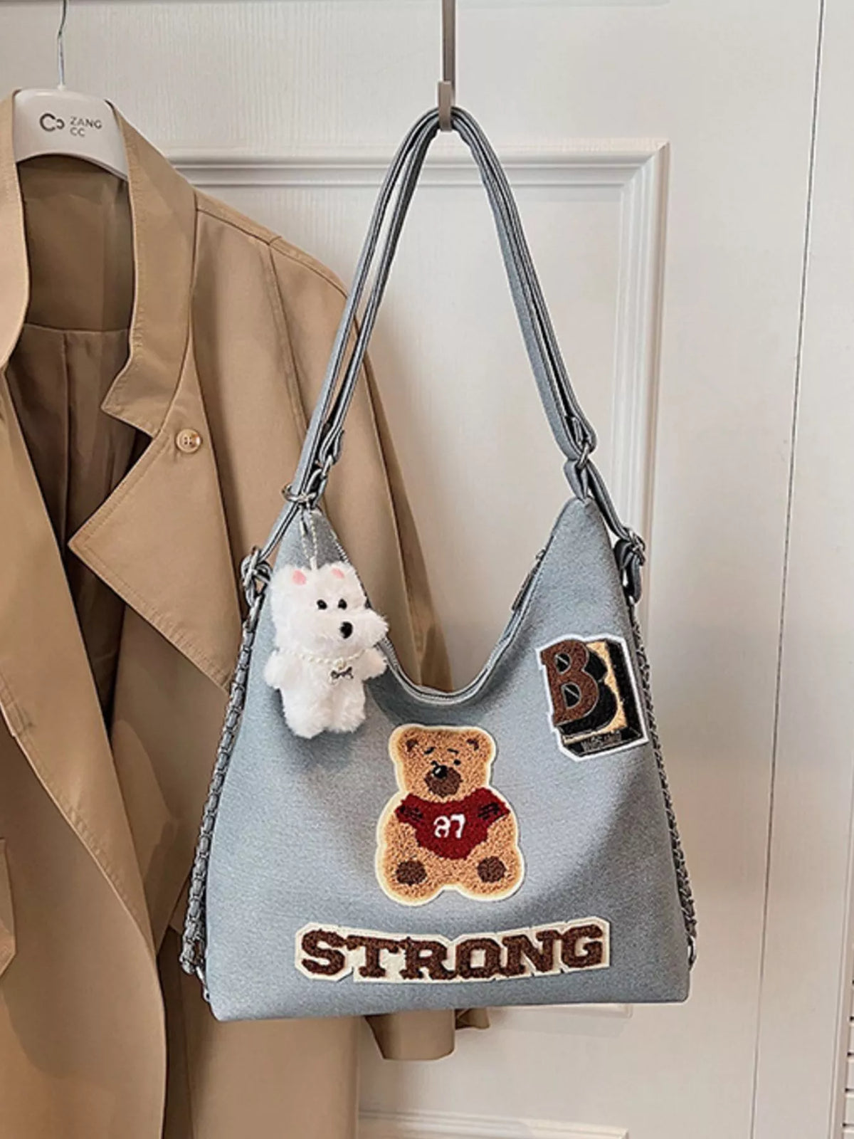 Gray handbag with patches and a teddy bear charm hanging on a hook against a white wall.