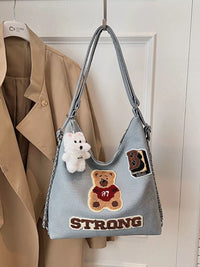Gray handbag with patches and a teddy bear charm hanging on a hook against a white wall.