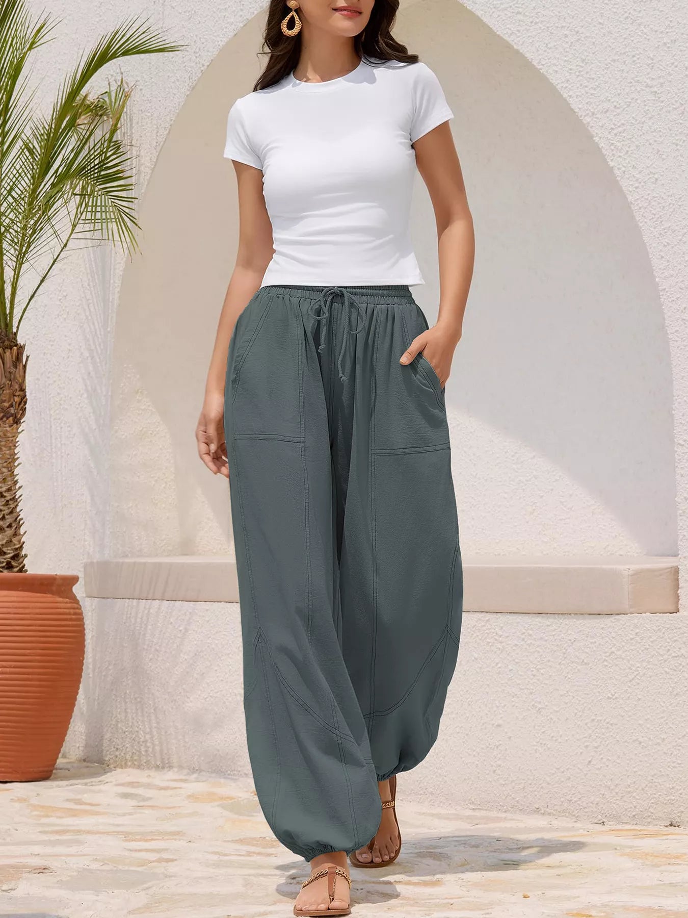 Woman wearing a white top and gray wide-leg pants standing in front of a stone wall with a plant.