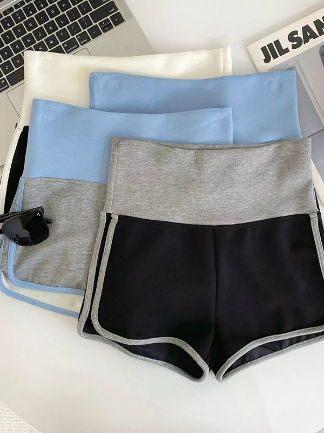 Set of black, gray, and blue shorts on a white surface with a laptop and keyboard in the background.