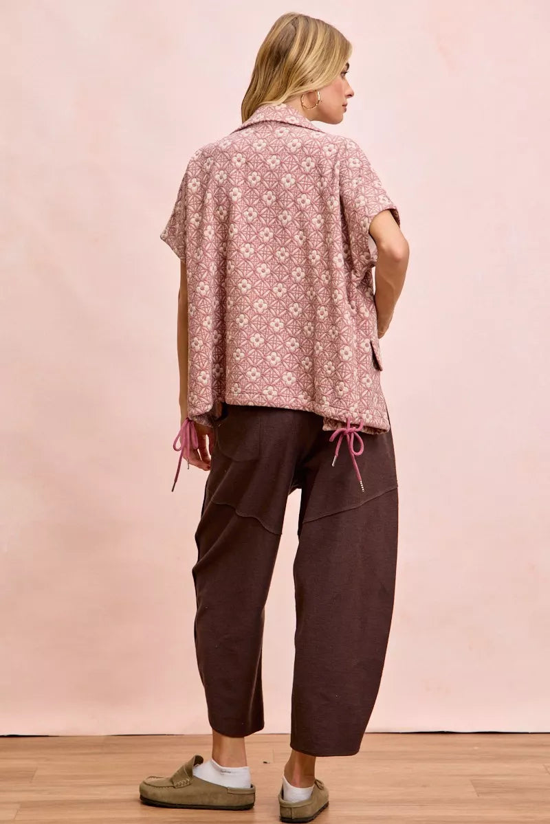 Woman wearing a patterned shirt and brown pants against a pink background