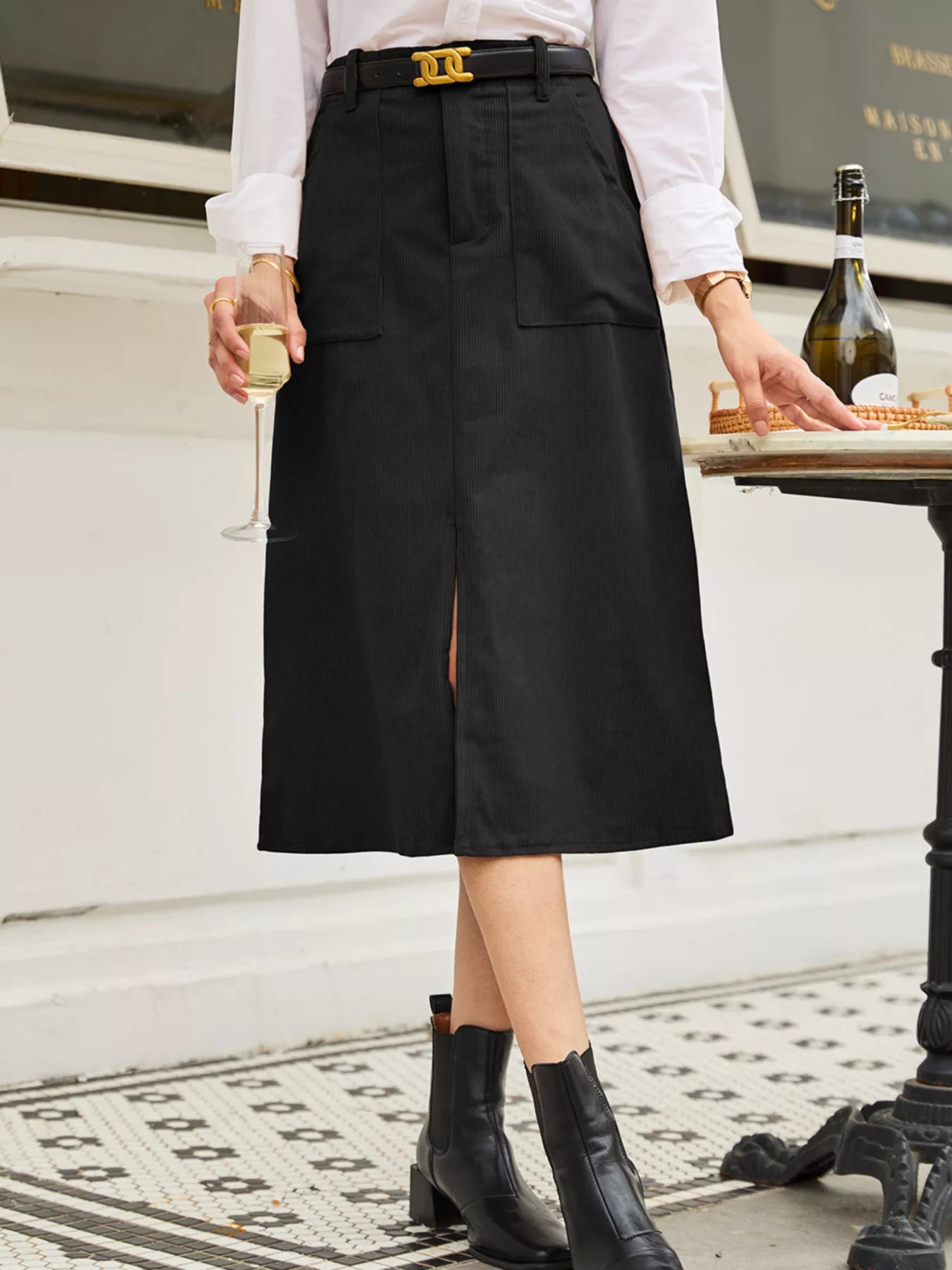 Person wearing a black skirt with a white shirt and black boots, holding a glass.