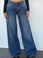 A person wearing loose-fit medium-wash blue denim jeans with a black long-sleeve top, standing against a plain light wall.
