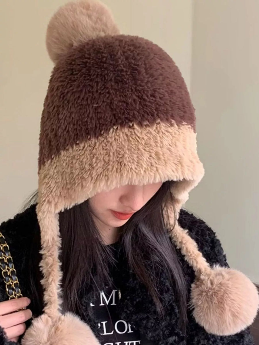 Person wearing a brown and beige fur-trimmed hat with pom-poms on a plain background