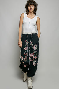 Woman wearing a white sleeveless top and black pants with floral embroidery on a gray background