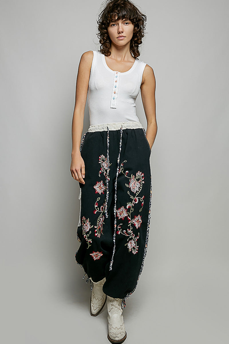 Woman wearing a white sleeveless top and black pants with floral embroidery on a gray background