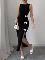 Woman wearing a black dress with white bow details, holding a small white handbag against a plain background.