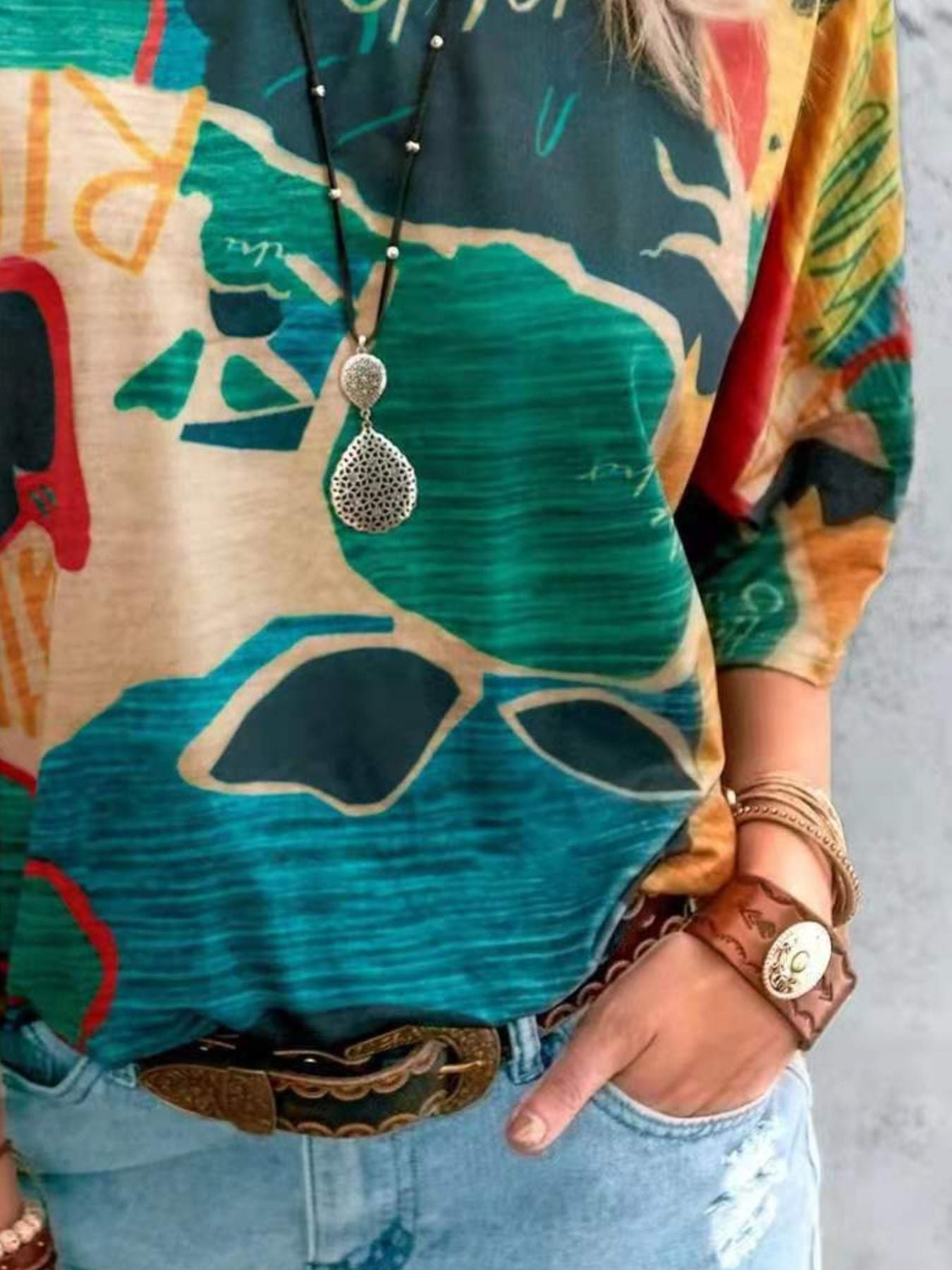 Person wearing a colorful patterned shirt with a necklace and bracelets, standing against a neutral background.