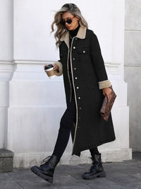 Woman wearing a long black coat with fur trim, holding a coffee cup and a phone, standing against a white wall.