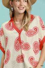 Woman wearing a cream blouse with red paisley patterns and a straw hat against a light blue background
