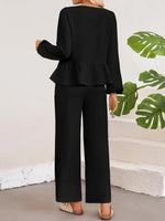 Person wearing a black peplum top and pants with a white wall and green leaves in the background