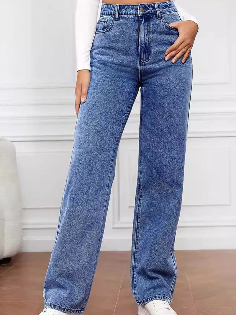 A person wearing high-waisted, wide-leg blue denim jeans with a white long-sleeve top and white sneakers, standing in front of a white paneled wall.