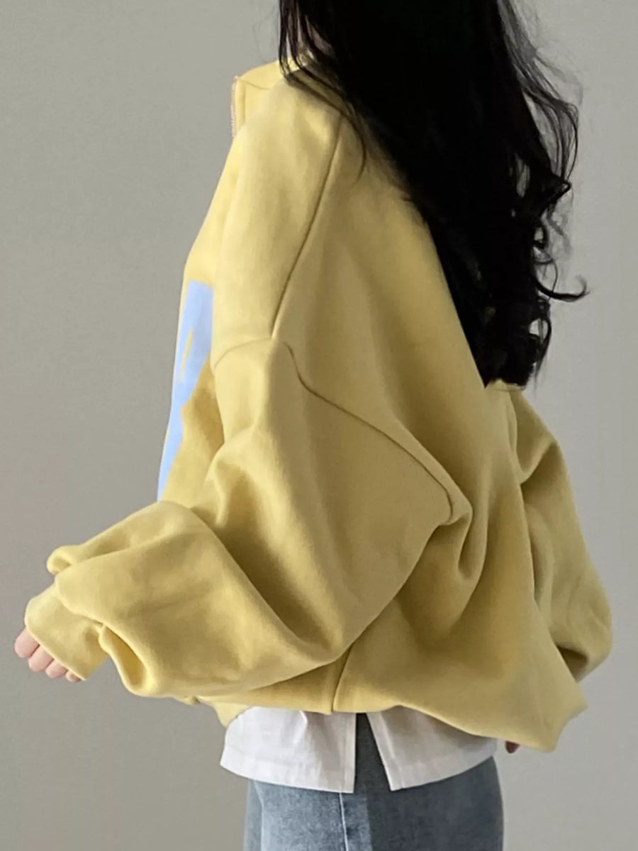 A person wearing an oversized pale yellow sweatshirt with a light blue graphic print on the sleeve, layered over a white shirt and blue jeans, shown from the back against a plain gray wall.