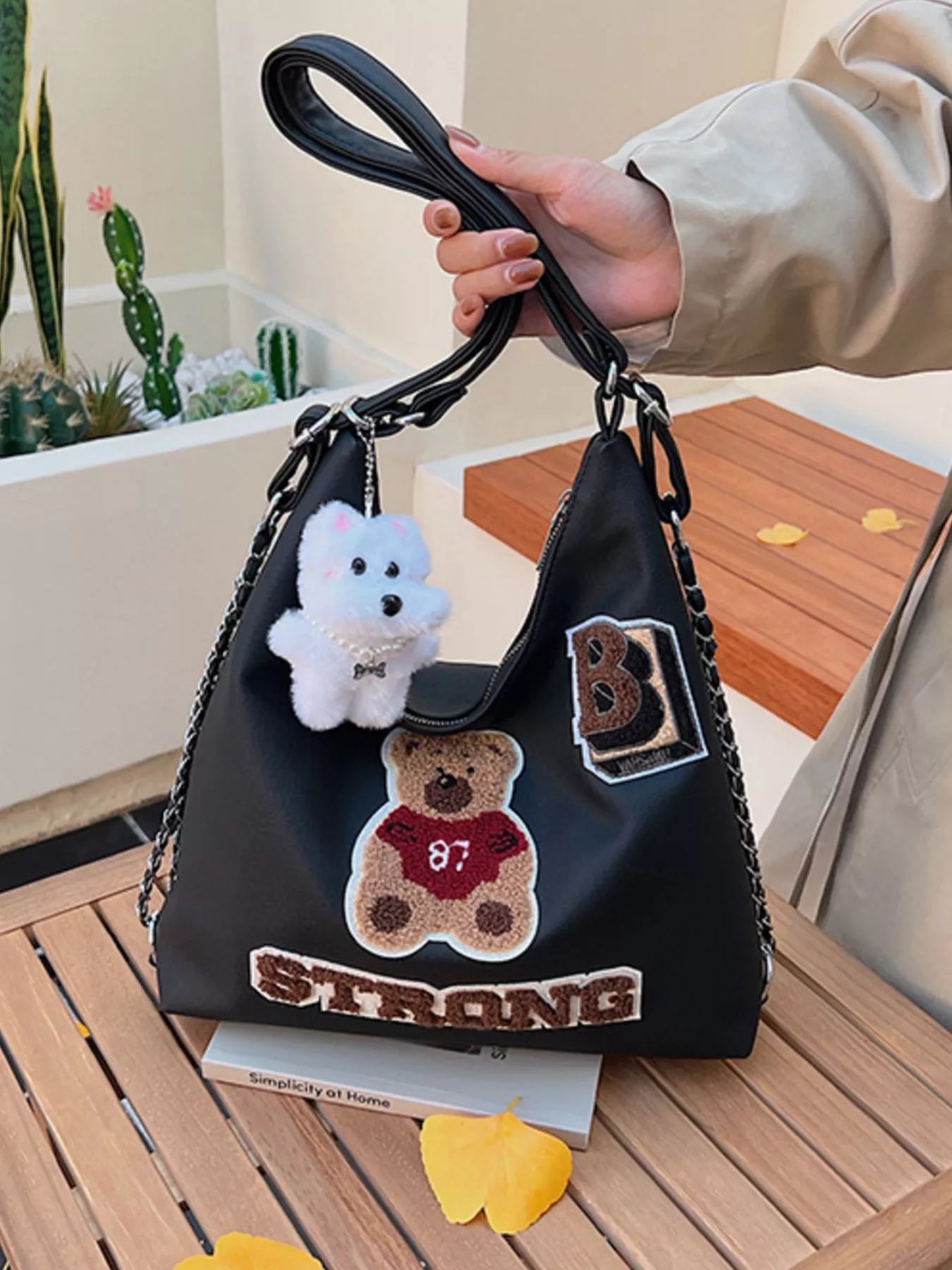 Black handbag with teddy bear and 'STRONG' patch held by a person outdoors.
