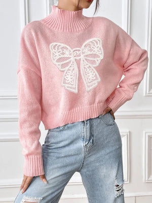 Pink sweater with a white lace bow design worn by a person against a white wall.