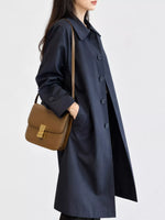 A woman wearing a long navy blue trench coat and carrying a brown leather shoulder bag with a gold clasp, photographed against a plain light gray studio background.