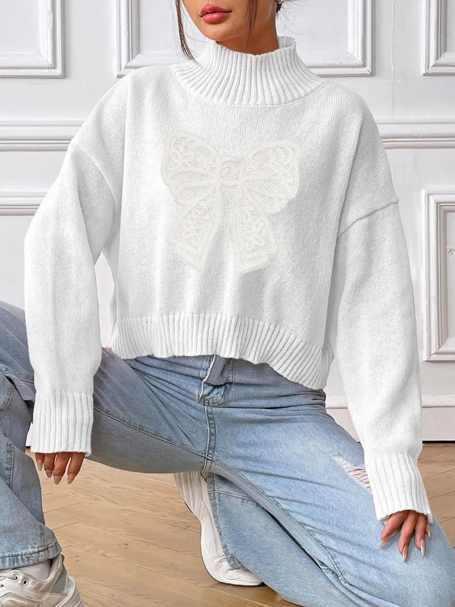 Person wearing a white turtleneck sweater and light blue jeans in a room with white walls.