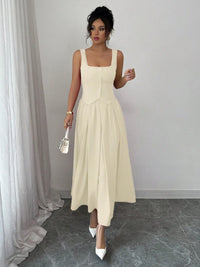 Woman wearing a beige sleeveless dress standing in a room with white curtains and gray floor.