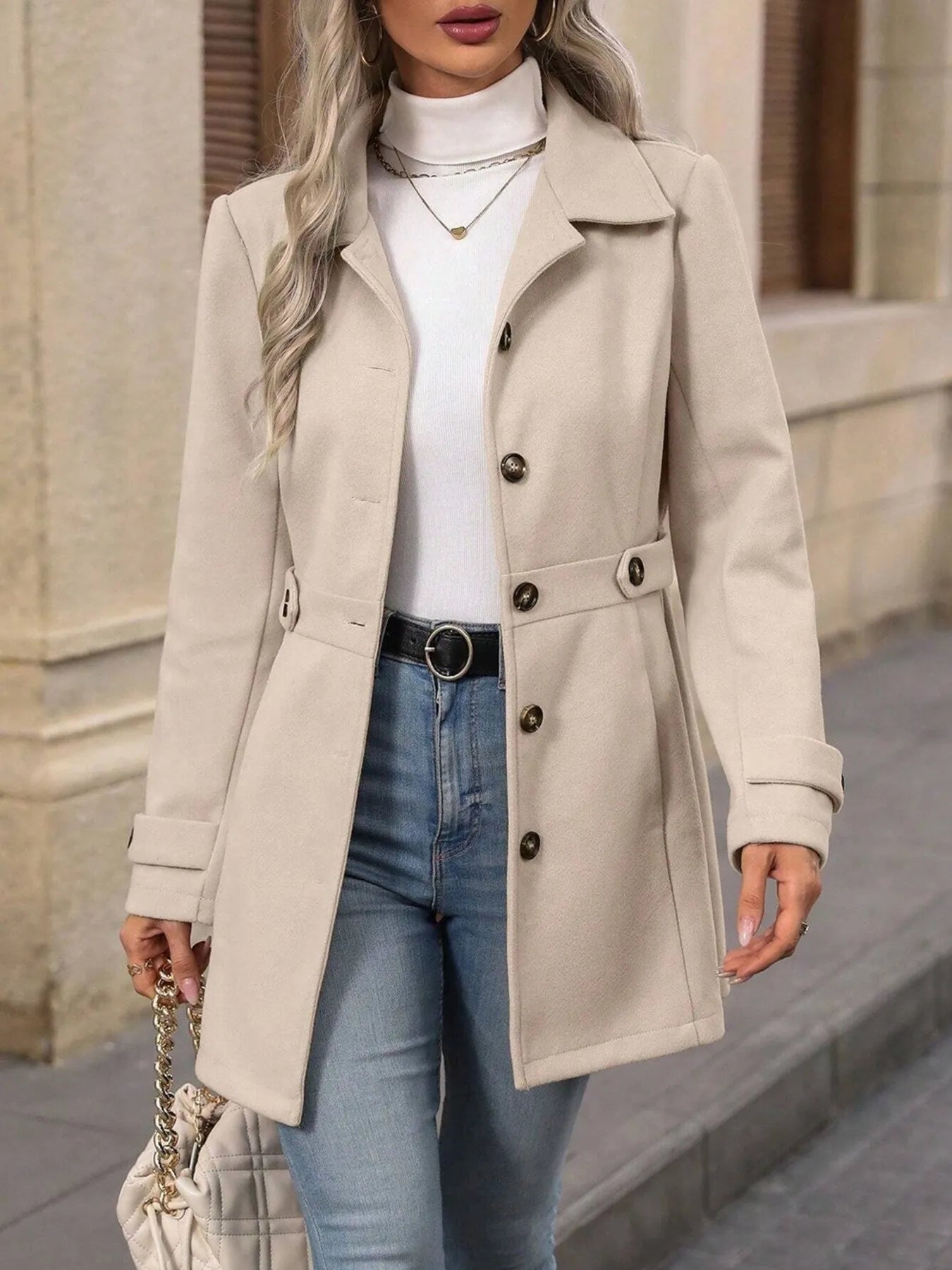 Woman wearing a beige coat, white turtleneck, and blue jeans with a neutral background