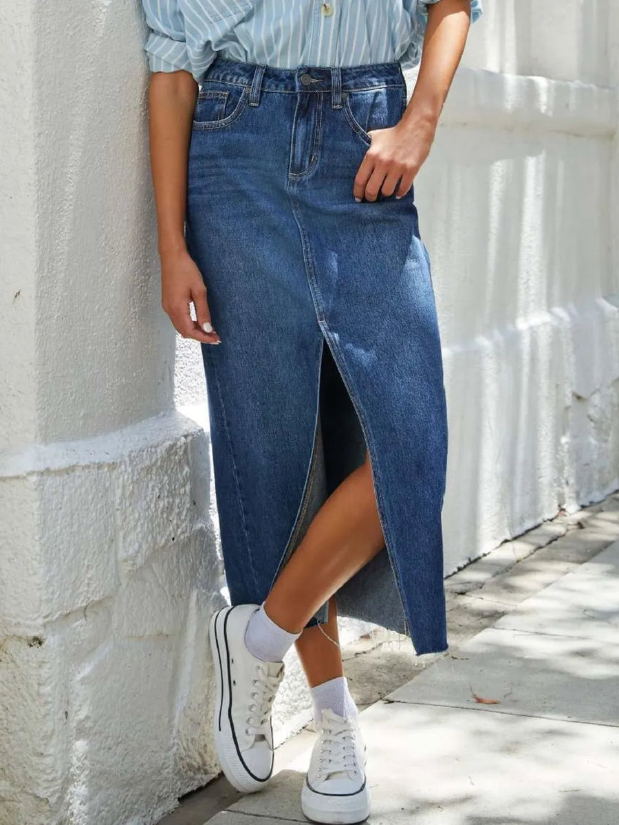 Person wearing a blue denim skirt with white sneakers against a light stone wall.