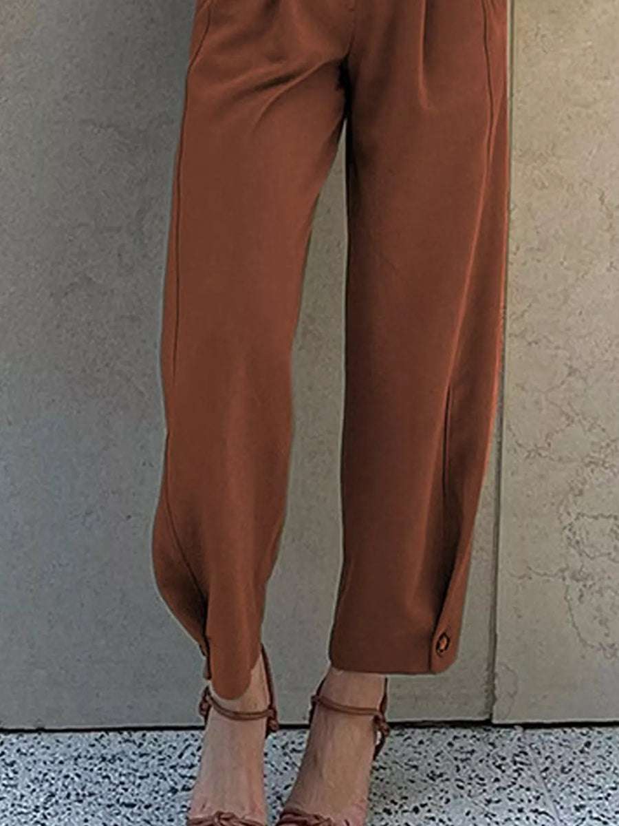 A person wearing rust-brown wide-leg cropped pants with side buttons at the hem, paired with brown strappy high-heeled sandals, standing against a textured concrete wall.